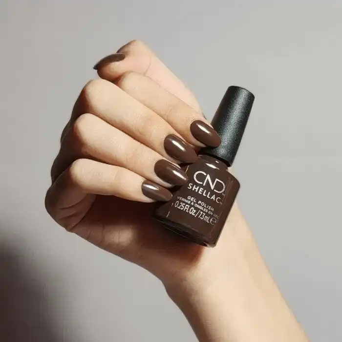 Hand holding CND Shellac polish bottle with matching manicured nails against gray background.
