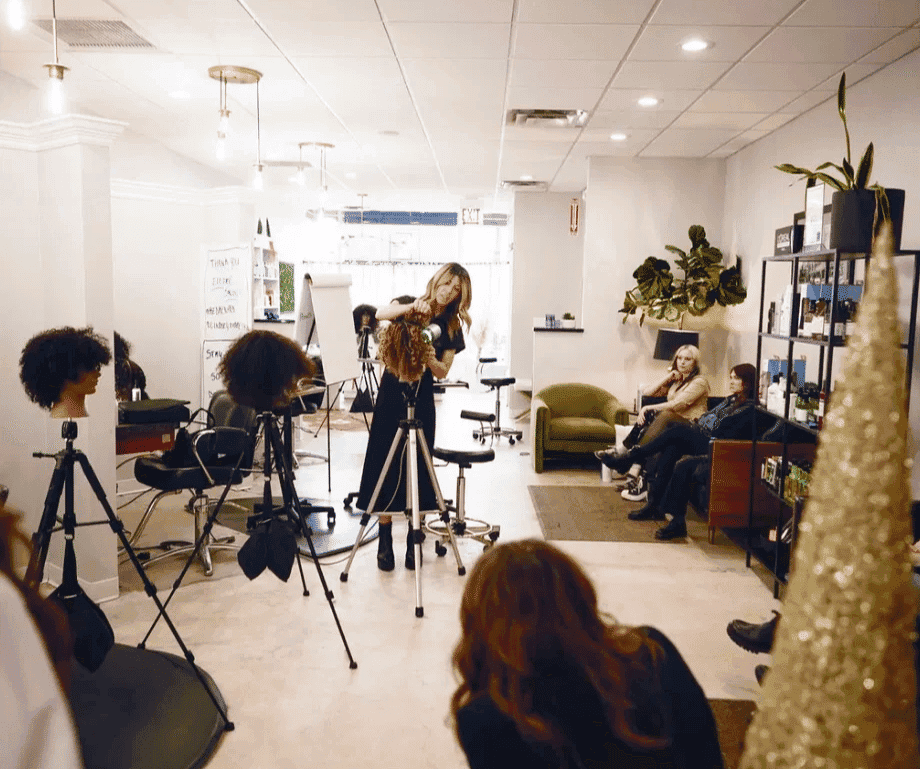 Hairdresser teaching a styling class in a salon setting with several attendees.